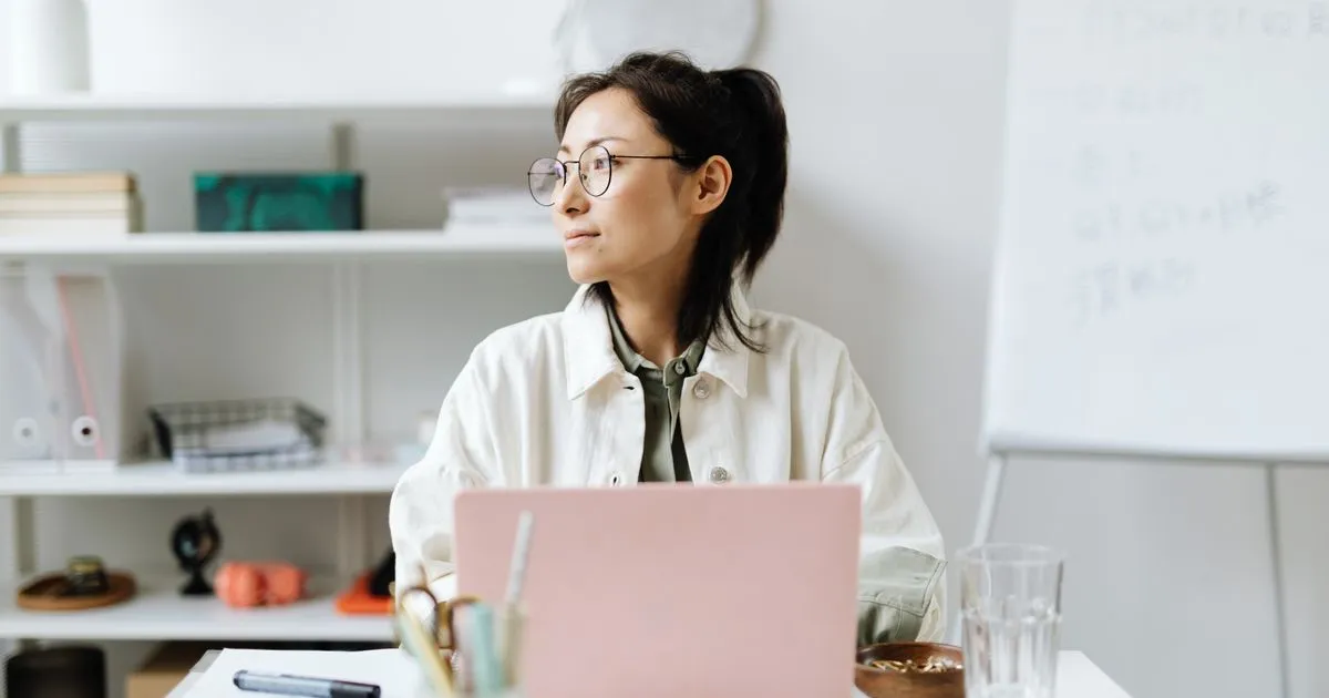 A woman with glasses sitting at a desk with a laptop, looking to the side