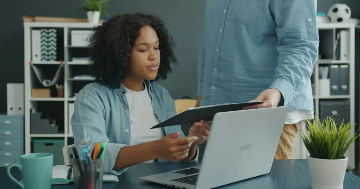 A colleague holding a clipboard and asking a seated coworker a question at her laptop