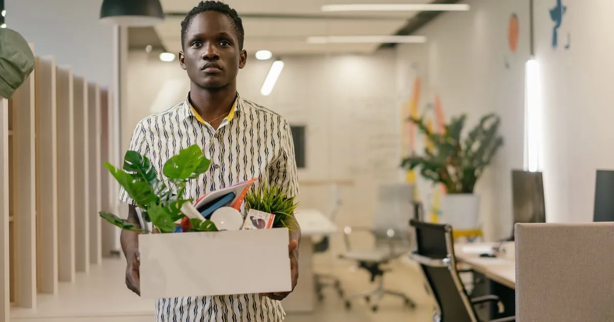 An employee carrying a box of belongings and a plant through an office